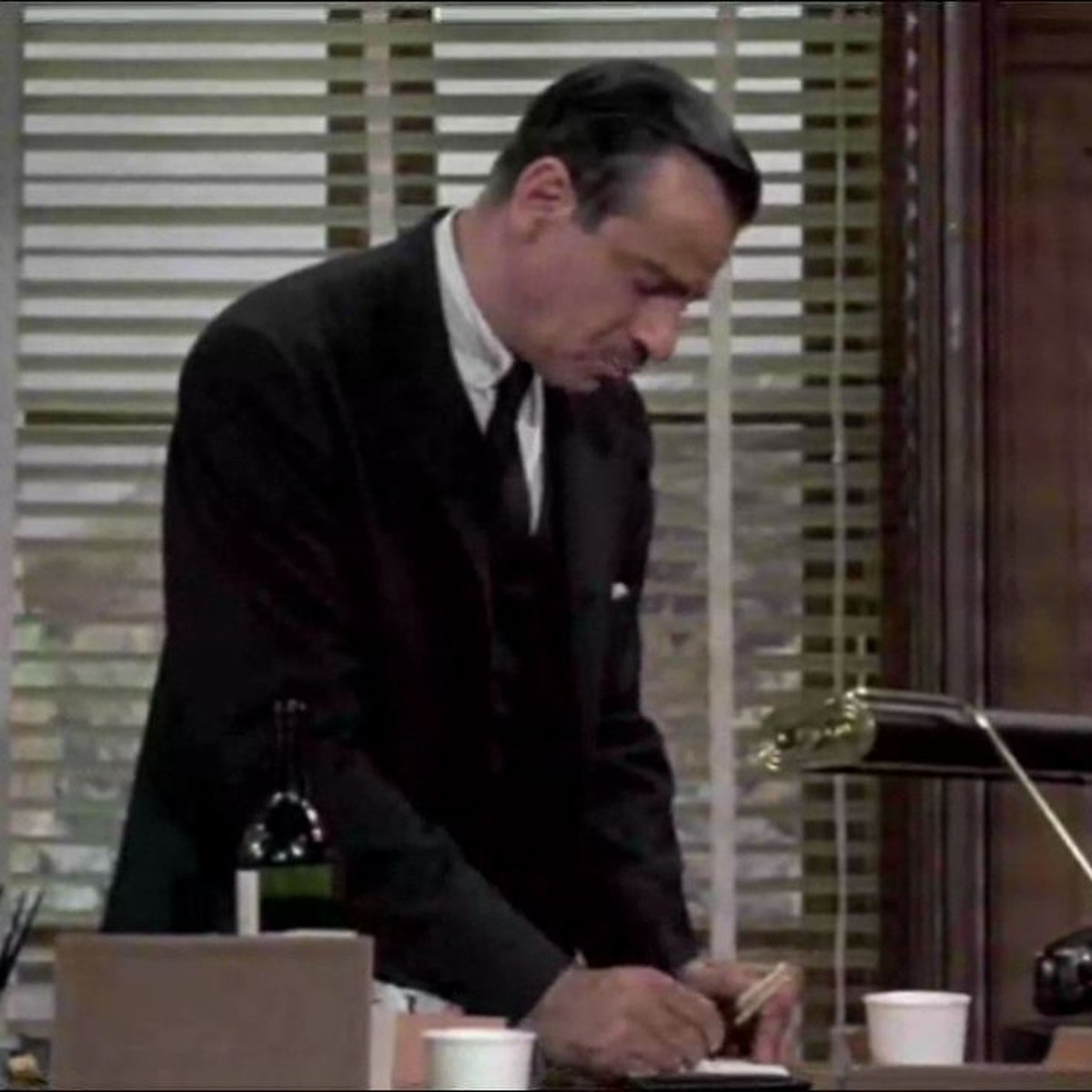 Still from Charade (1963): a man working at a desk of papers and phones